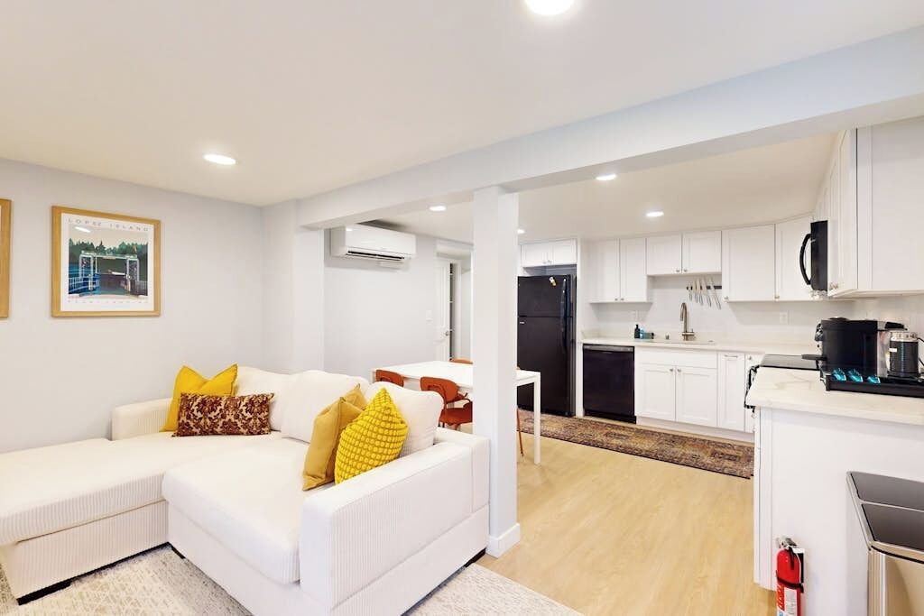 A modern living room with a white sofa, a dining area, and a kitchen with white cabinets and a black refrigerator.