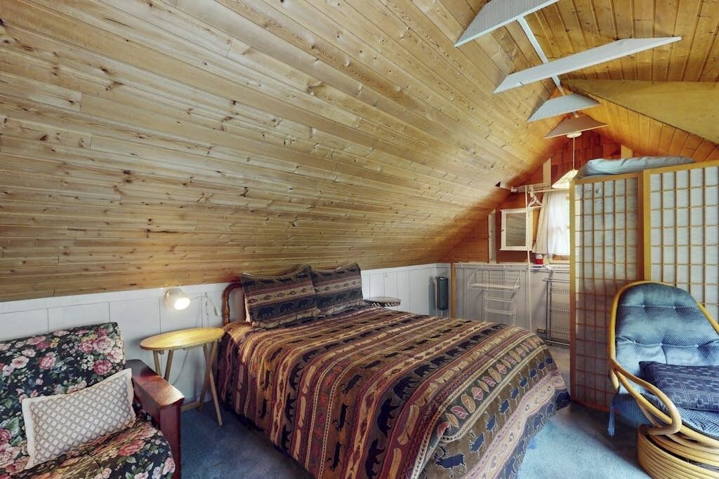 A room with a sloped ceiling, a bed with patterned bedding, a chair, and a small table.