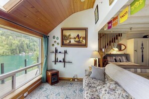 Quarto com teto de madeira, cama com cabeceira, sofá, mesa pequena, luminária, pintura e janela com vista para um lago.