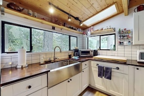 Une cuisine moderne avec des armoires blanches, des appareils en acier inoxydable et une grande fenêtre offrant une vue sur la forêt.
