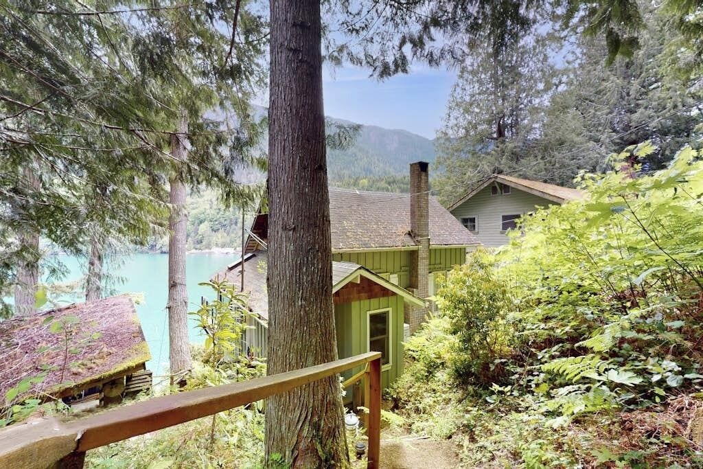 A cabin with a wooden deck, surrounded by dense forest and a lake.