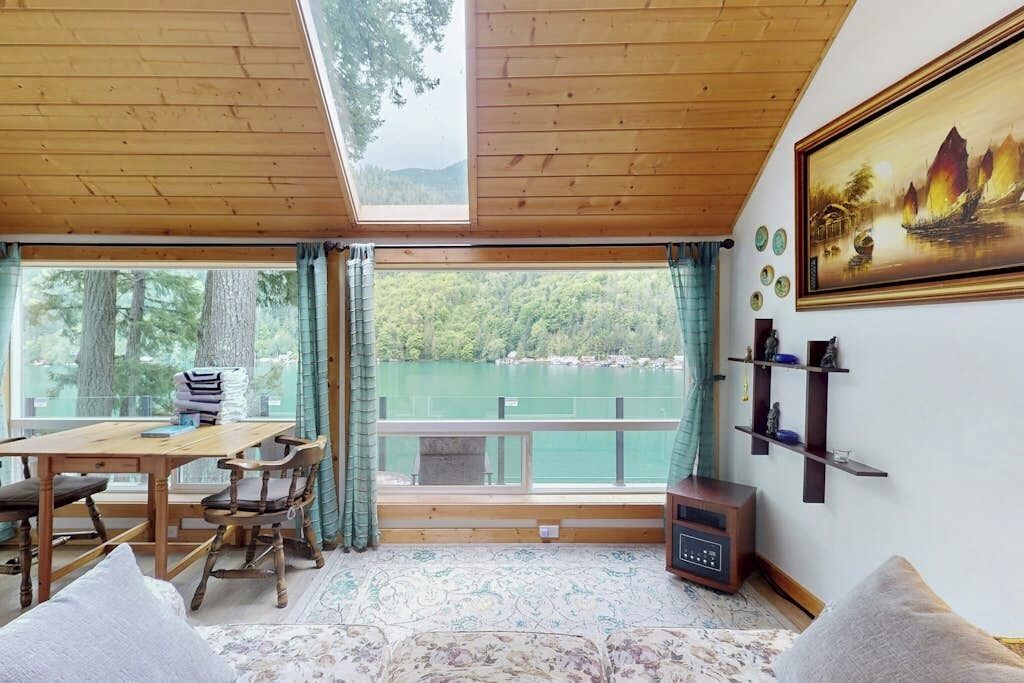 A room with a wooden ceiling, a table with chairs, a sofa, a painting, and a view of a lake.