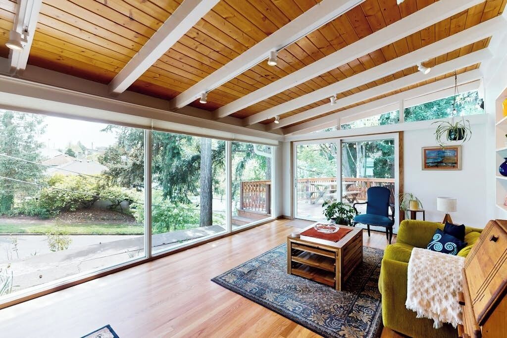 Une pièce spacieuse avec de grandes fenêtres, des poutres en bois au plafond, un canapé vert, un fauteuil bleu et une petite table surmontée d’un tapis.