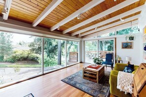 A spacious room with large windows, wooden beams on the ceiling, a green sofa, a blue chair, and a small table with a rug. - 1BR Home- Tranquil Mid-Century retreat (Seattle)