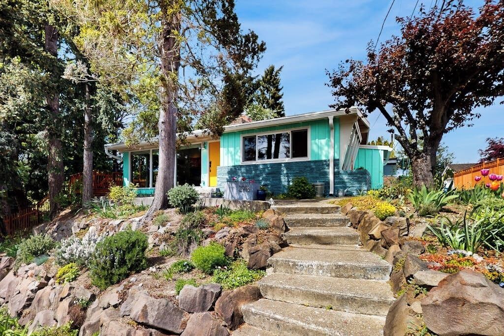 A two-story house with a stone staircase leading to the front door, surrounded by a garden with various plants and trees.