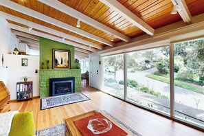 A spacious living room with a fireplace, wooden ceiling beams, a bookshelf, and a large window offering a view of the outdoors. - 1BR Home- Tranquil Mid-Century retreat (Seattle)