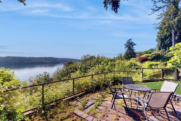 Une terrasse avec des sièges extérieurs donnant sur un lac, entourée d’une végétation luxuriante et sous un ciel dégagé.