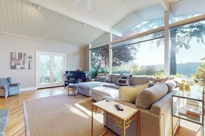 A spacious living room with a piano, a sectional sofa, and a glass coffee table. - 2BR Lakefront home with deck & garage parking (Lake Forest Park)