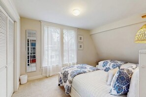 Quarto com duas camas, janela com cortinas leves, luminária de teto e abajur fixado na parede.