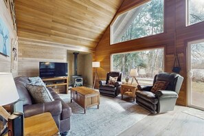 Una sala de estar acogedora con chimenea, sofás de cuero, un televisor de pantalla plana y amplios ventanales con vistas al bosque.