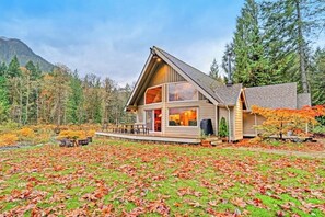 A cabin with a large deck, surrounded by a forest and a mountain backdrop. - Upgraded 3BR Riverfront - Hot Tub - WoodStove (Sultan)