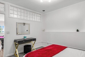 Ein Schlafzimmer mit einem Bett, einem Nachttisch, einem Spiegel, einem Fenster mit Jalousien und einem kleinen Regal.