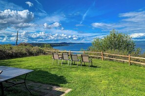 A grassy outdoor area with a wooden fence, a table, and chairs facing a body of water. - 2BR Vashon Island Home with Breathtaking views (Vashon)