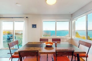 Zona de dinar amb taula gran i cadires, un bol, i vista al mar a través de grans finestres.