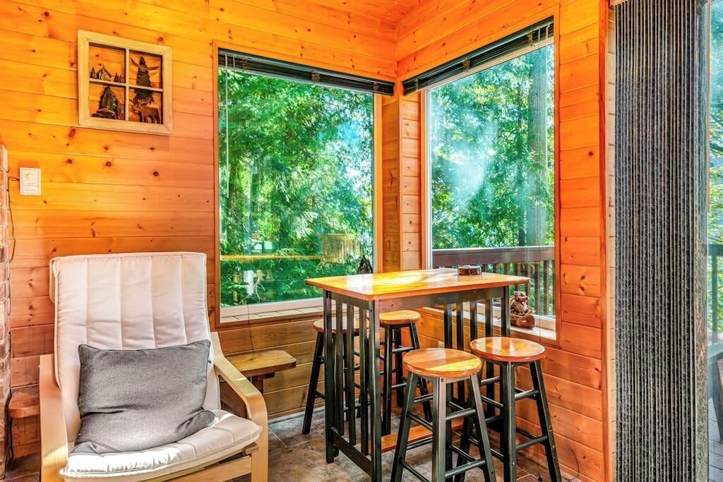 A wooden interior with a window view of greenery, a cushioned chair, a wooden table with stools, and a small decorative item on the table.