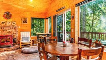 A wooden cabin with a fireplace, a dining table, and a deck overlooking a forest.