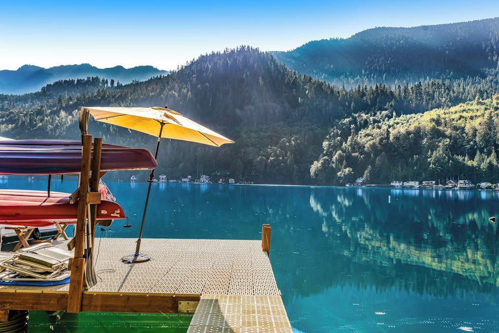 A dock with a boat and an umbrella, a calm lake, and forested hills in the background.