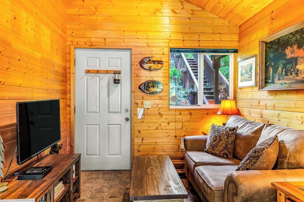 Ein gemütliches Blockhaushaus-Interieur mit Fernseher, Holzwänden, einer Couch und einem Fenster mit Blick auf eine Holzterrasse.