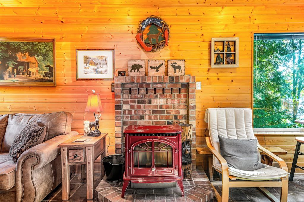 A cozy cabin interior with a brick fireplace, wooden walls, a leather sofa, and a window with a view of trees.