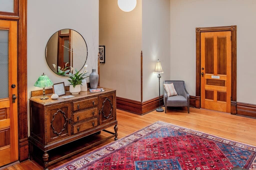A wooden dresser with drawers, a round mirror, a green lamp, and a red patterned rug in a room with wooden flooring and a door.