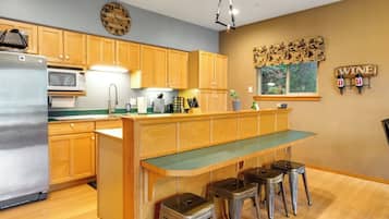 A kitchen with wooden cabinets, a stainless steel refrigerator, a microwave, a sink, and a bar area with stools.