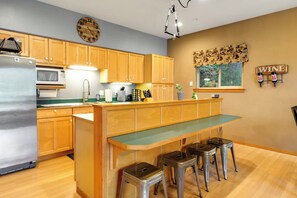 A kitchen with wooden cabinets, a stainless steel refrigerator, a microwave, a sink, and a bar area with stools.