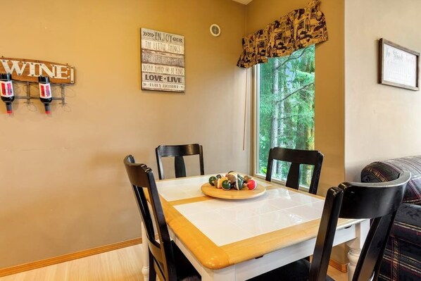 A dining area with a table set for four, chairs, a wine rack with bottles, and a window with a view of trees.