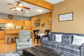A living room with a sofa, chairs, a coffee table, a ceiling fan, and a kitchen area with wooden cabinets and a microwave.