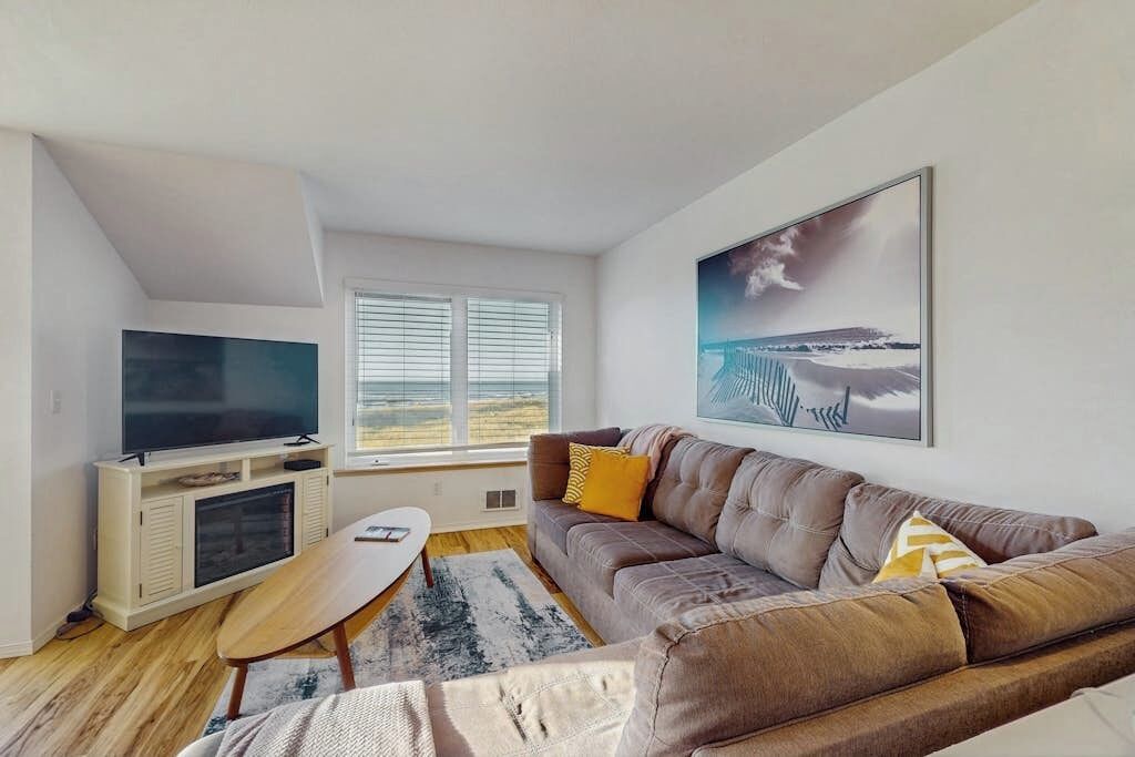 Ein modernes Wohnzimmer mit einem Flachbildfernseher, einem Sofa, einem Couchtisch und einem großen Fenster mit Jalousien.