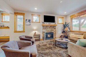 A living room with a stone fireplace, a television mounted above it, and a large window with wooden frames. - BlackBearLodge 4BR with ensuites - walk everywhere (Cle Elum)