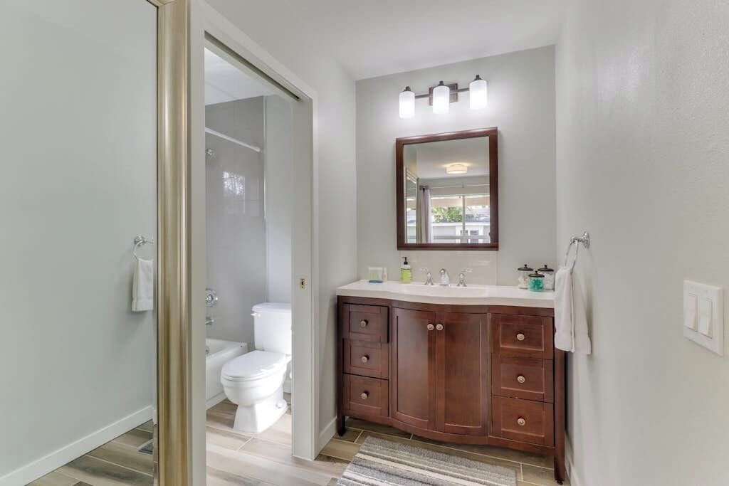 Un baño con inodoro blanco, tocador de madera con espejo y zona de ducha.