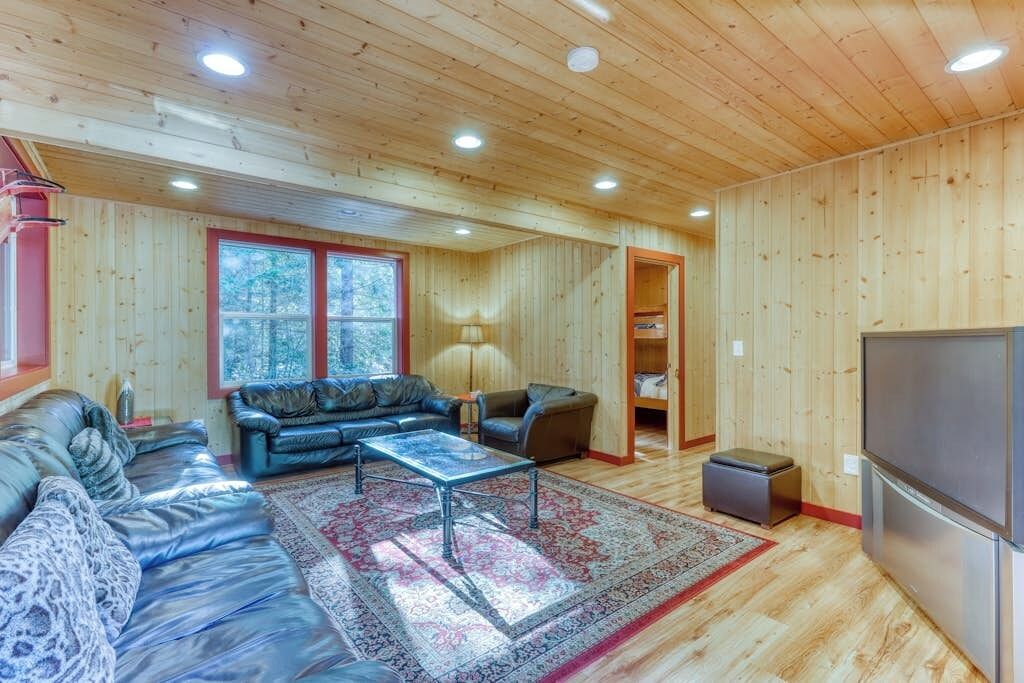 Un salón amplio con un sofá grande, una mesa de centro y una alfombra. El techo de madera tiene iluminación empotrada y una ventana que ofrece vistas al exterior.