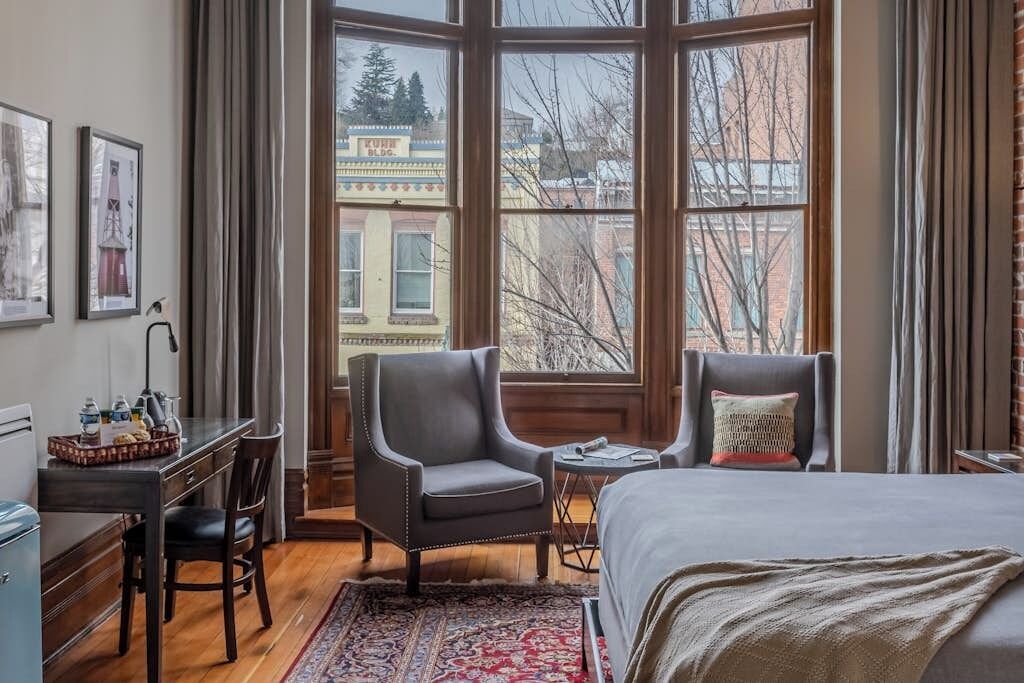 A hotel room with a large window, a bed, a desk with a chair, and a small table.
