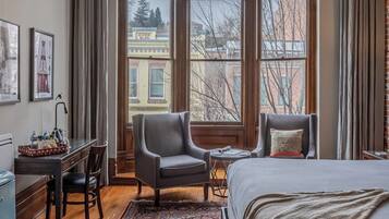 A hotel room with a large window, a bed, a desk with a chair, and a small table.