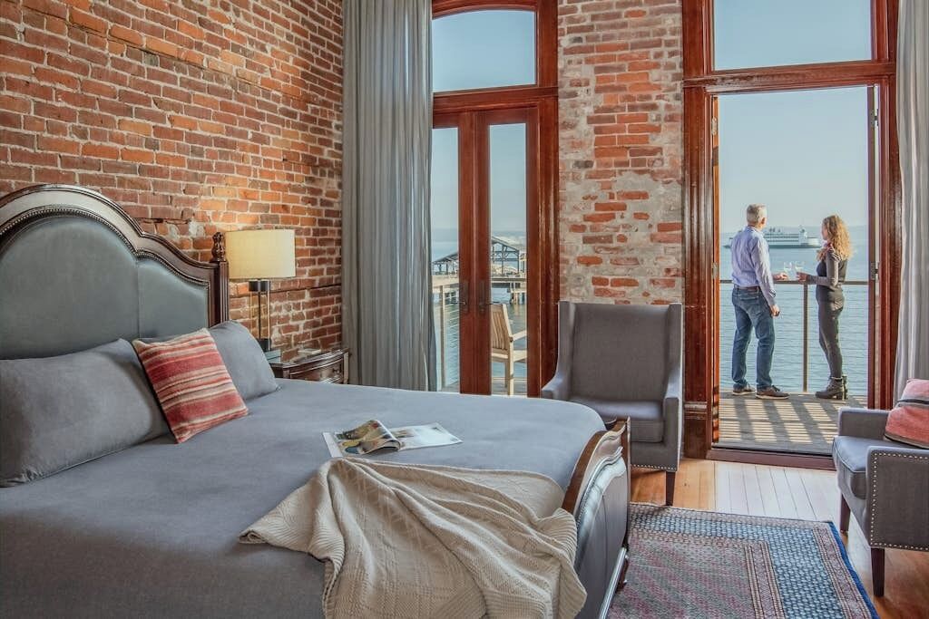 A hotel room with a large bed, a chair, a table, and a view of the water through a large window.