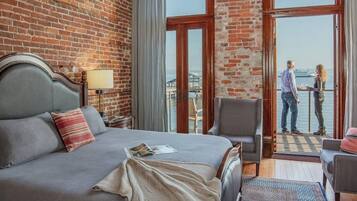 A hotel room with a large bed, a chair, a table, and a view of the water through a large window.