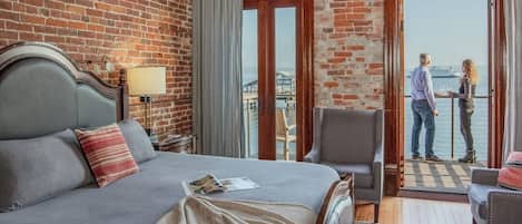 A hotel room with a large bed, a chair, a table, and a view of the water through a large window.