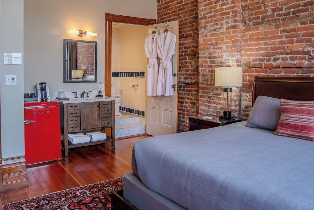 Hotelkamer met een bed, een rode minibars kastje, een houten dressoir, een badkamer met douche en een bakstenen muur.