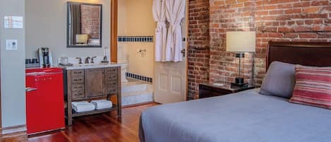 A hotel room with a bed, a red minibar, a wooden dresser, a bathroom with a shower, and a brick wall.