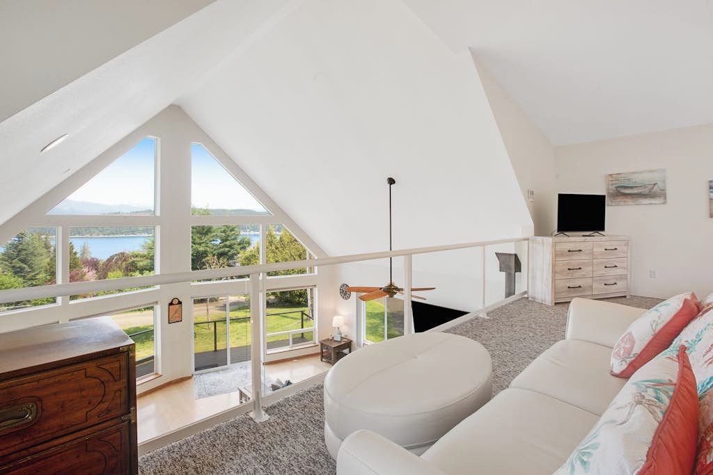 Una habitación con techos altos, techo blanco, un sofá blanco, una televisión y vistas a un paisaje verde a través de amplios ventanales.