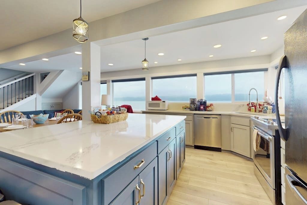 A modern kitchen with a large island, stainless steel appliances, and a view through the windows.
