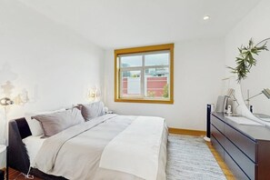 Ein Schlafzimmer mit einem Bett, Nachttisch, Schminktisch und einem Fenster mit Blick auf Gebäude.
