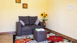 Ein Wohnzimmer mit einem grauen Sofa, einem kleinen Hocker, einer Vase mit Sonnenblumen und einem blumigen Teppich.