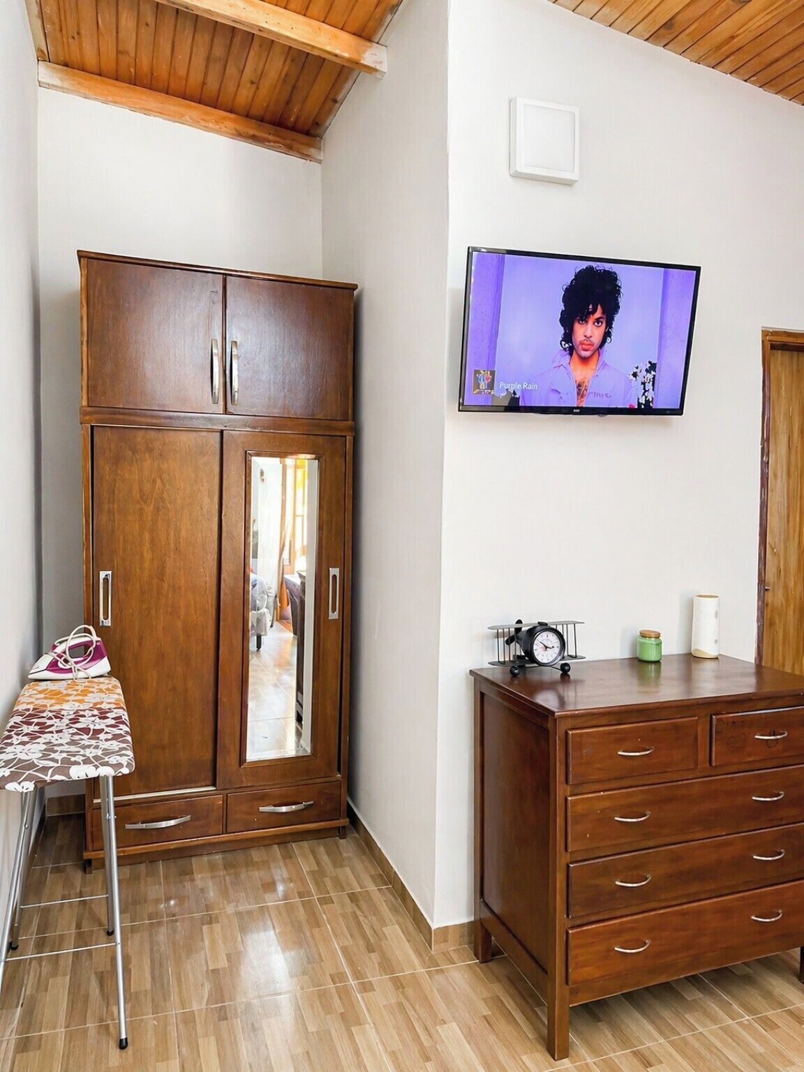 Ein Zimmer mit einem Holzschrank, einem an der Wand befestigten Fernseher und einem kleinen Tisch mit einer Handtasche darauf.