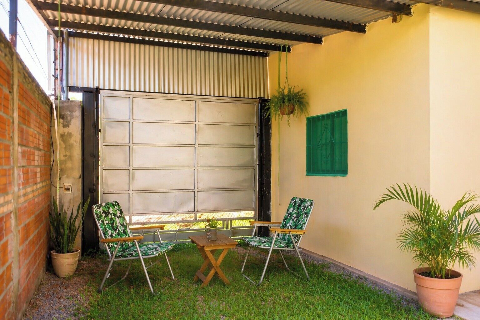 Un patio con techo de metal, pared de ladrillo y un persiano verde. Se observan dos sillas y una pequeña mesa.