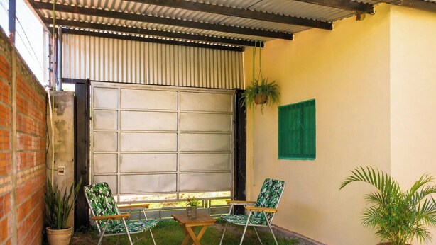 A patio area with a metal roof, a brick wall, and a green window shutter. There are two chairs and a small table.