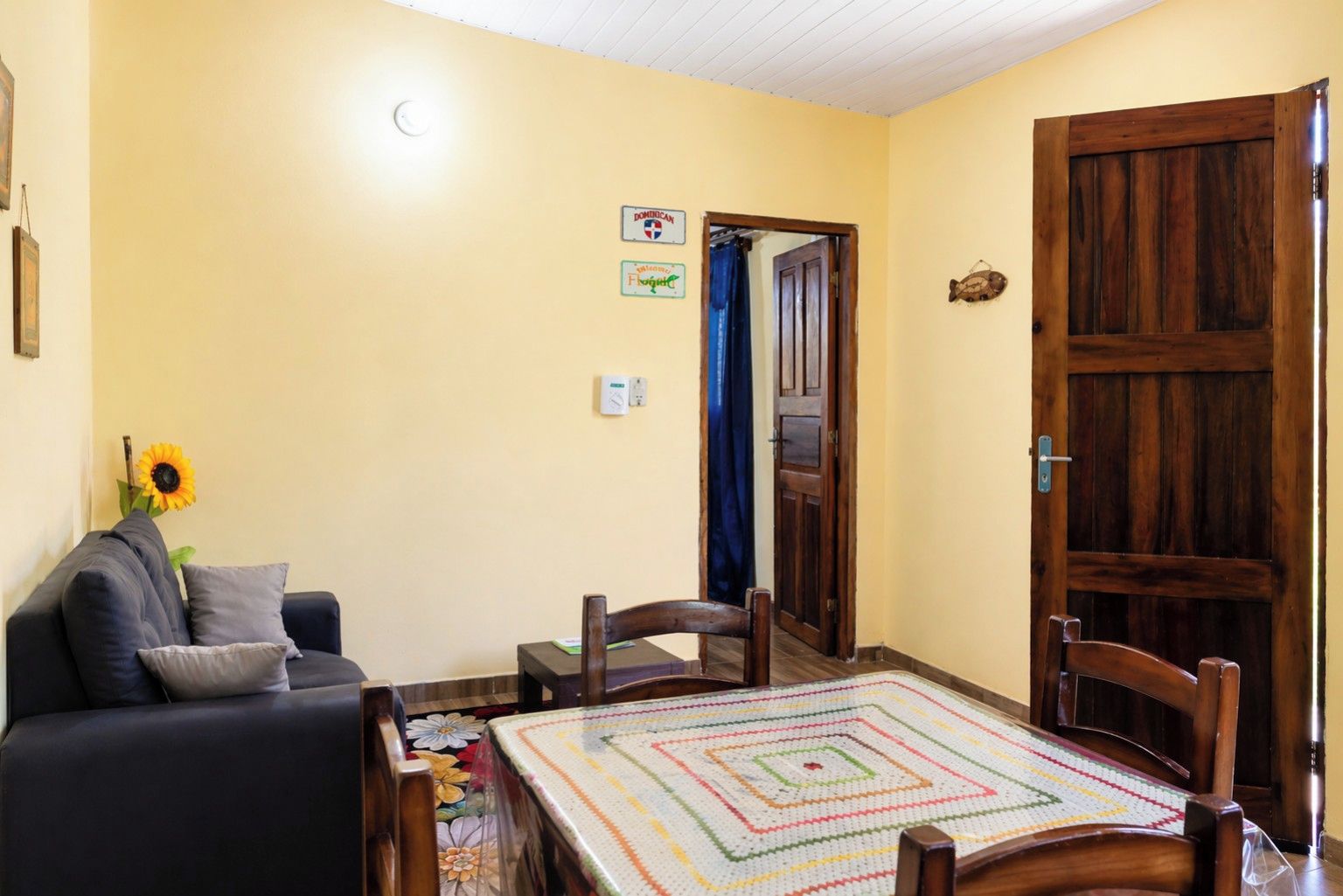 Una sala de estar acogedora con un sofá, una mesa de centro con un mantel colorido, una puerta de madera y un termostato fijado a la pared.