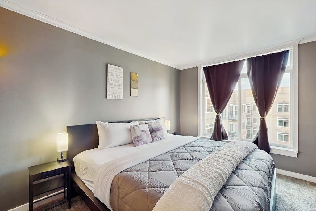 A hotel room with a bed, a nightstand, a window with curtains, and a wall with two framed pictures.