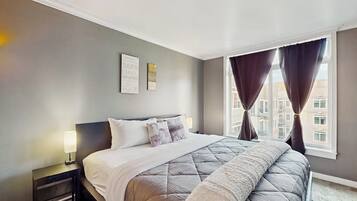 A hotel room with a bed, a nightstand, a window with curtains, and a wall with two framed pictures.