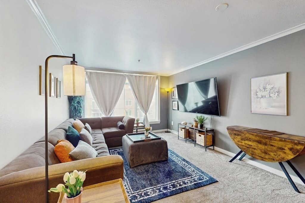 Un salon moderne comprenant un canapé, un pouf, une table basse, un téléviseur et une petite table d’appoint en bois.